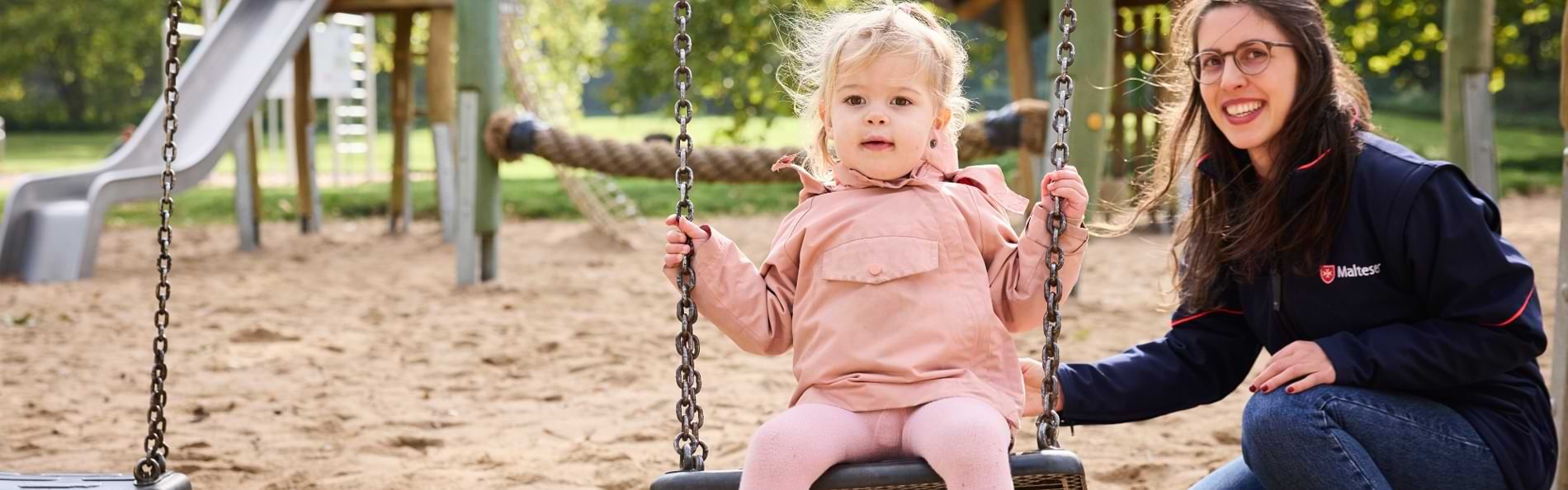 Ein Kleinkind in rosa Kleidung sitzt auf einer Schaukel auf einem Spielplatz. Neben ihm kniet eine erwachsene Frau mit Brille und dunkler Jacke und hält die Schaukel fest. Im Hintergrund sind Sandboden, Spielgeräte aus Holz, eine Rutsche und grüne Bäume zu sehen.