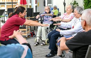 Ältere Menschen sitzen draußen in einer Reihe und strecken die Arme nach vorne als Übung