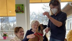 Ein Hund sitz auf dem Schoß einer älteren Frau, daneben eine Malteser Helferin und eine weitere Frau.