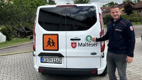 Ein Mann in Malteserkleidung klebt einen Aufkleber auf einen Malteser-Kleinbus.