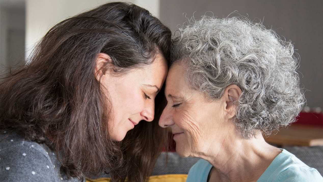 Eine junge Frau und eine ältere Frau lächeln und legen mit geschlossenen Augen ihre Köpfe aneinander. 