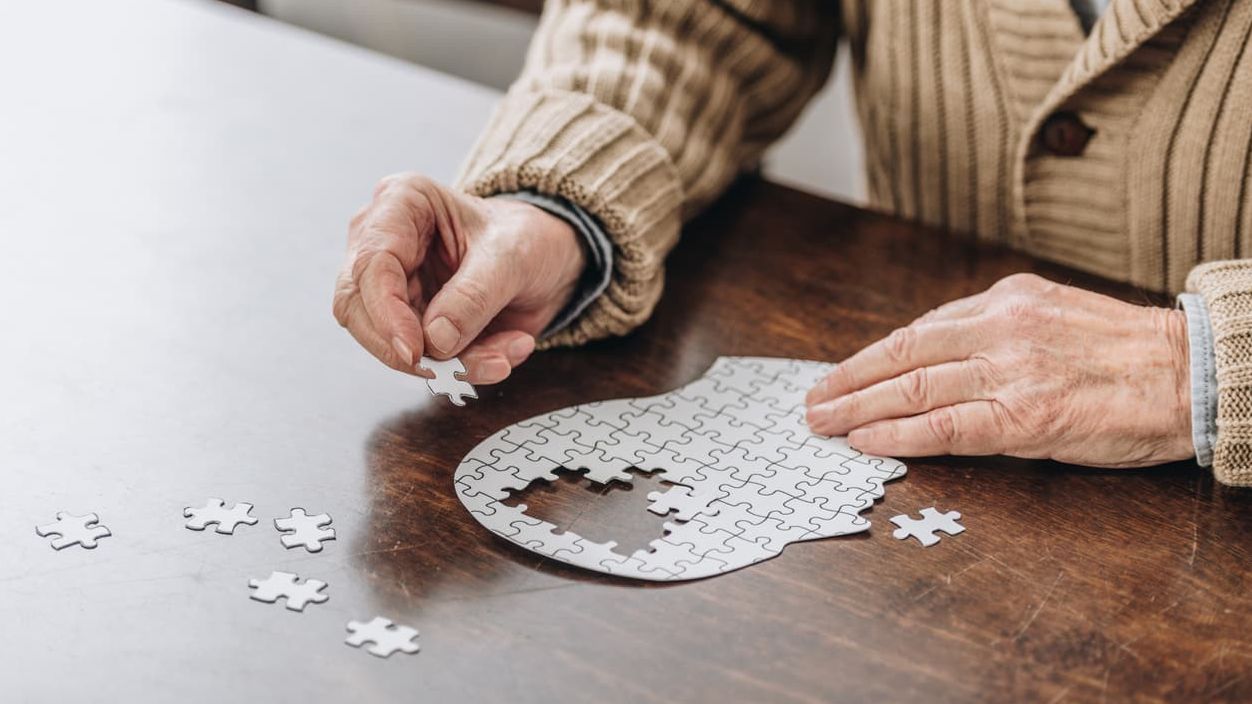 Älterer Mensch sitzt an einem Holztisch und setzt ein kopfförmiges Puzzle zusammen.