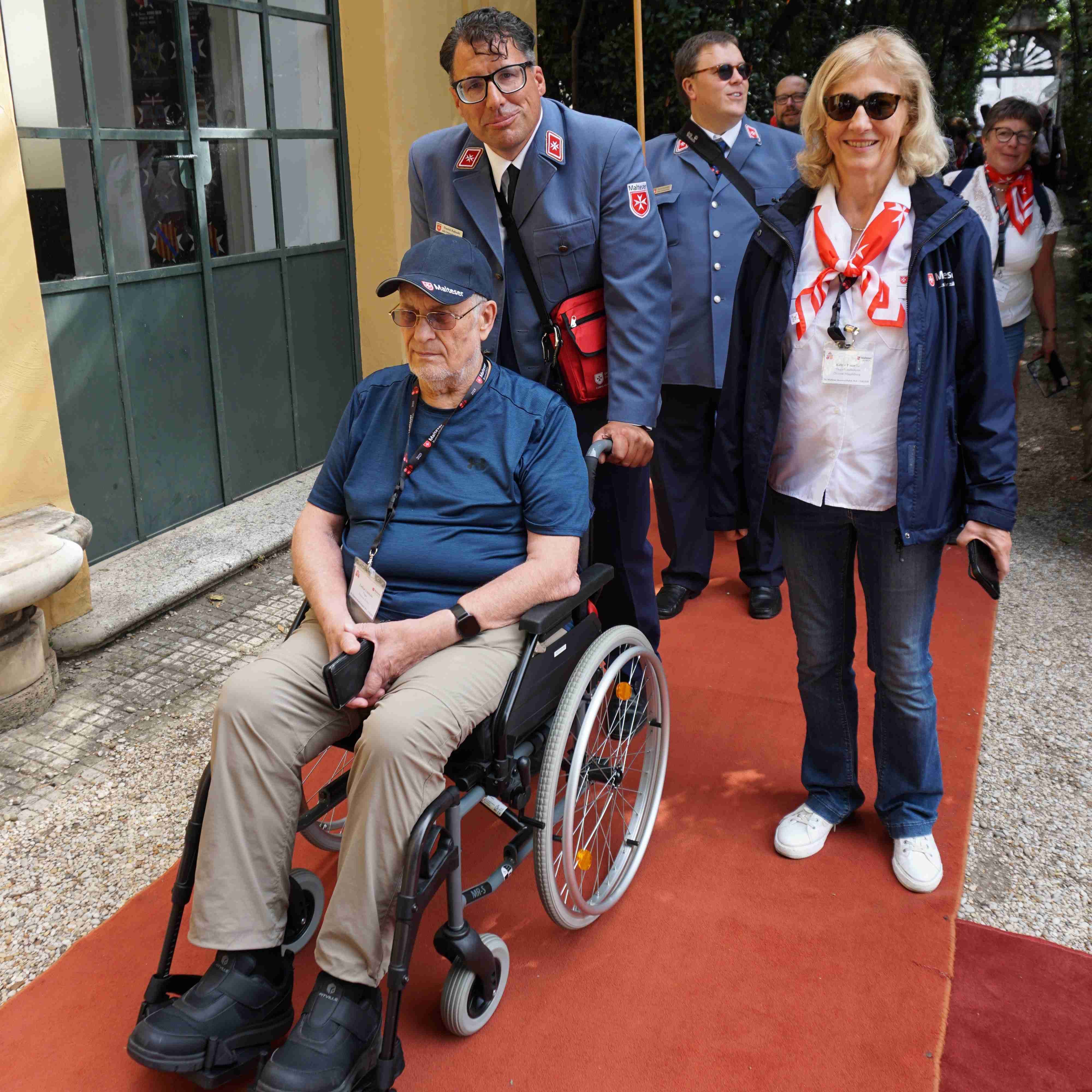 Person im Rollstuhl auf rotem Teppich, umgeben von Personen in blauer Uniform mit roten Halstüchern, vor einem Gebäude mit großen Fenstern.