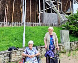 Eine Frau in einem Rollstuhl und eine Frau mit einer Gehhilfe stehen vor einem Gradierwerk.