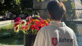 Eine Frau mit Malteser-Shirt (von hinten) und Blumen steht an einem Brunnen in Bad Reichenhall.
