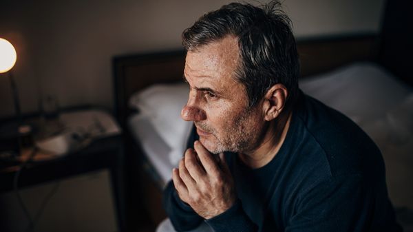 Ein älterer Mann sitzt in der Dunkelheit, nachdenklich auf seinem Bett.