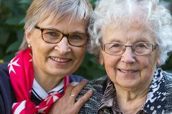 Malteser Mitarbeiterin legt Arm um ältere Frau und beide lachen in die Kamera.