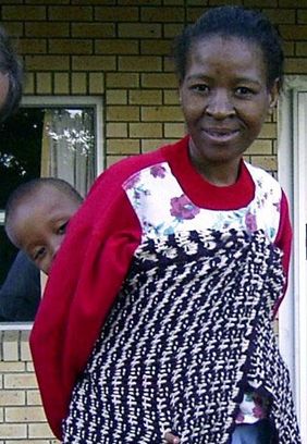 A woman with a child on her back stands in front of a brick wall.