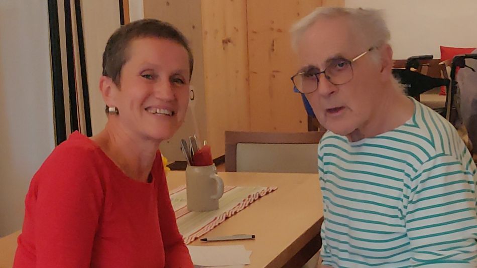 Eine lächelnde Frau mit kurzen Haaren und ein älterer Mann mit Brille an einem Tisch. 