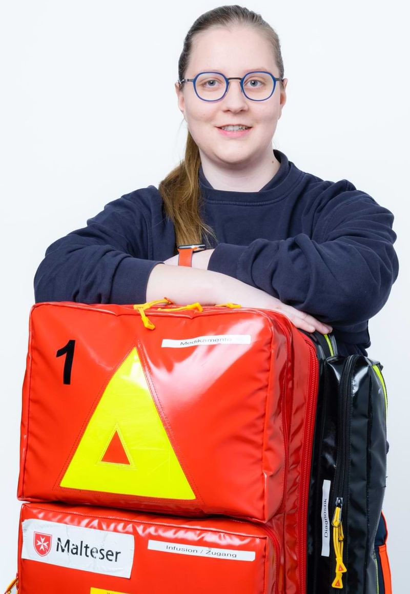 Eine junge Frau mit Brille hat ihre Arme auf einem Einsatzrucksack der Malteser SEG abgelegt.