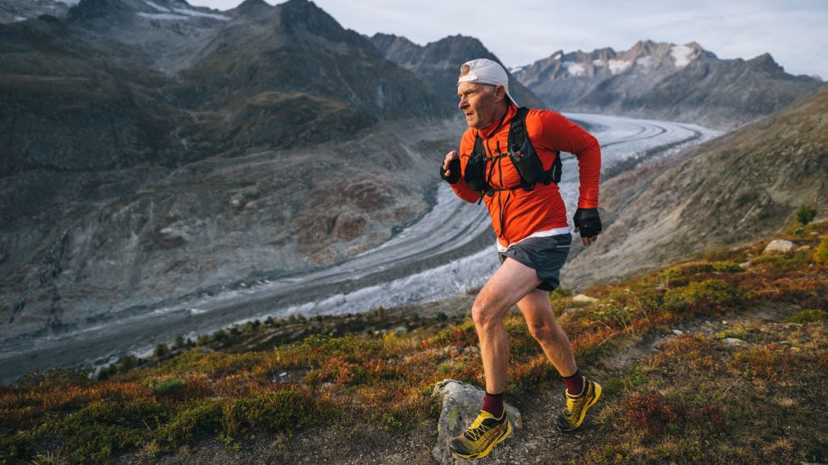 Ein älterer Mann in Laufkleidung bei einem Trailrun vor Bergkulisse. 