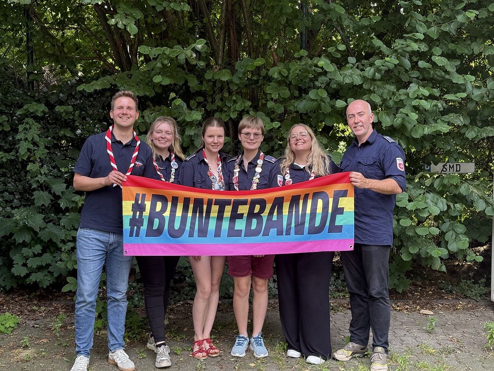 Der neue Diözesanführungskreis vor einer grünen Hecke mit einem Regenbogenbanner in den Händen, auf dem Hashtag Bunte Bande steht.