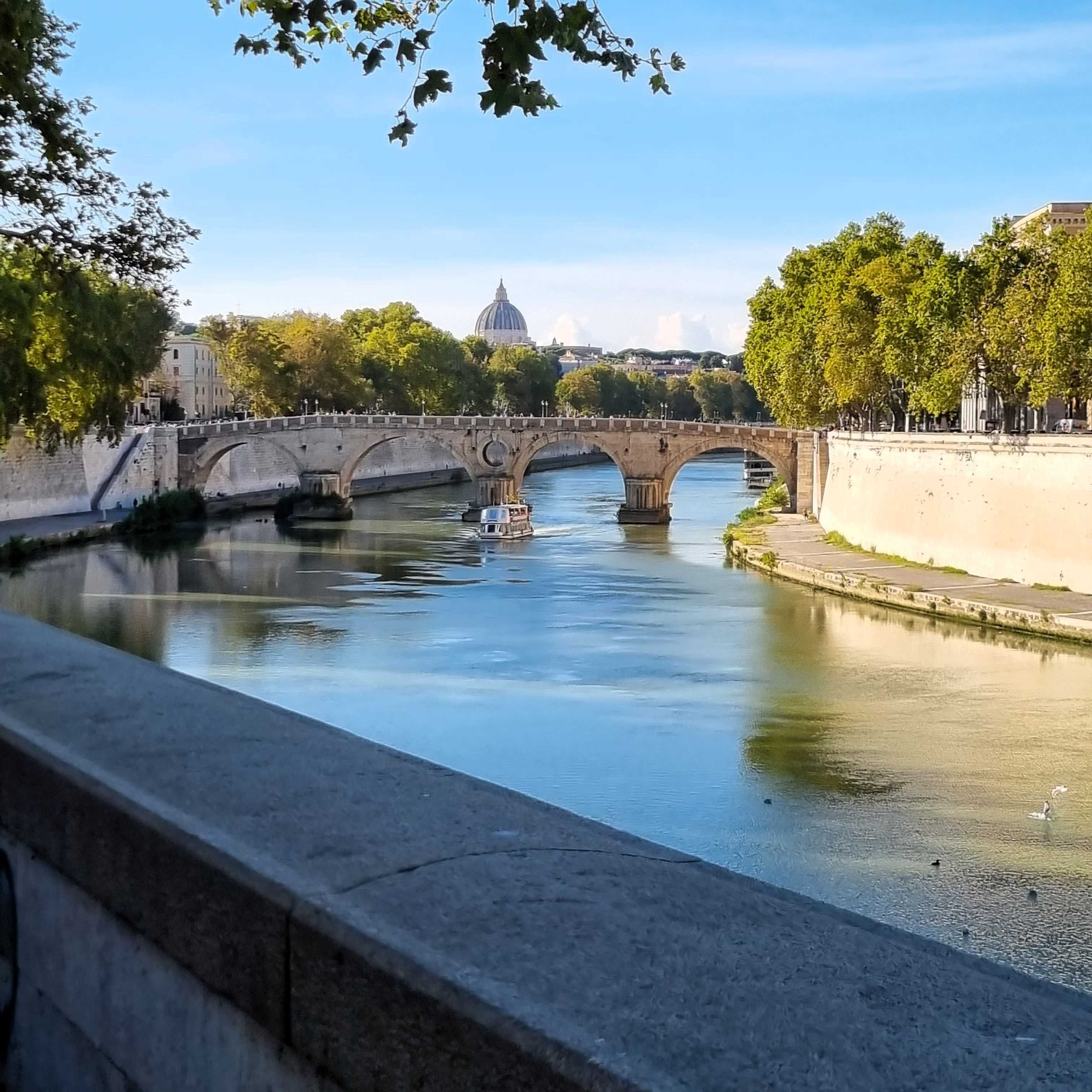 Fluss mit Steinbrücke und Bäumen an beiden Ufern unter blauem Himmel.