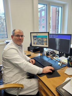 Arzt im weißen Kittel sitzt am Schreibtisch und betrachtet Röntgen- und 3D-Bilder auf zwei Monitoren.