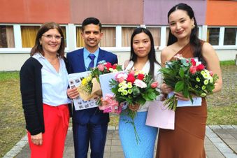 Vier Personen posieren mit Blumensträußen und Geschenken vor einem Gebäude – drei von ihnen elegant gekleidet.