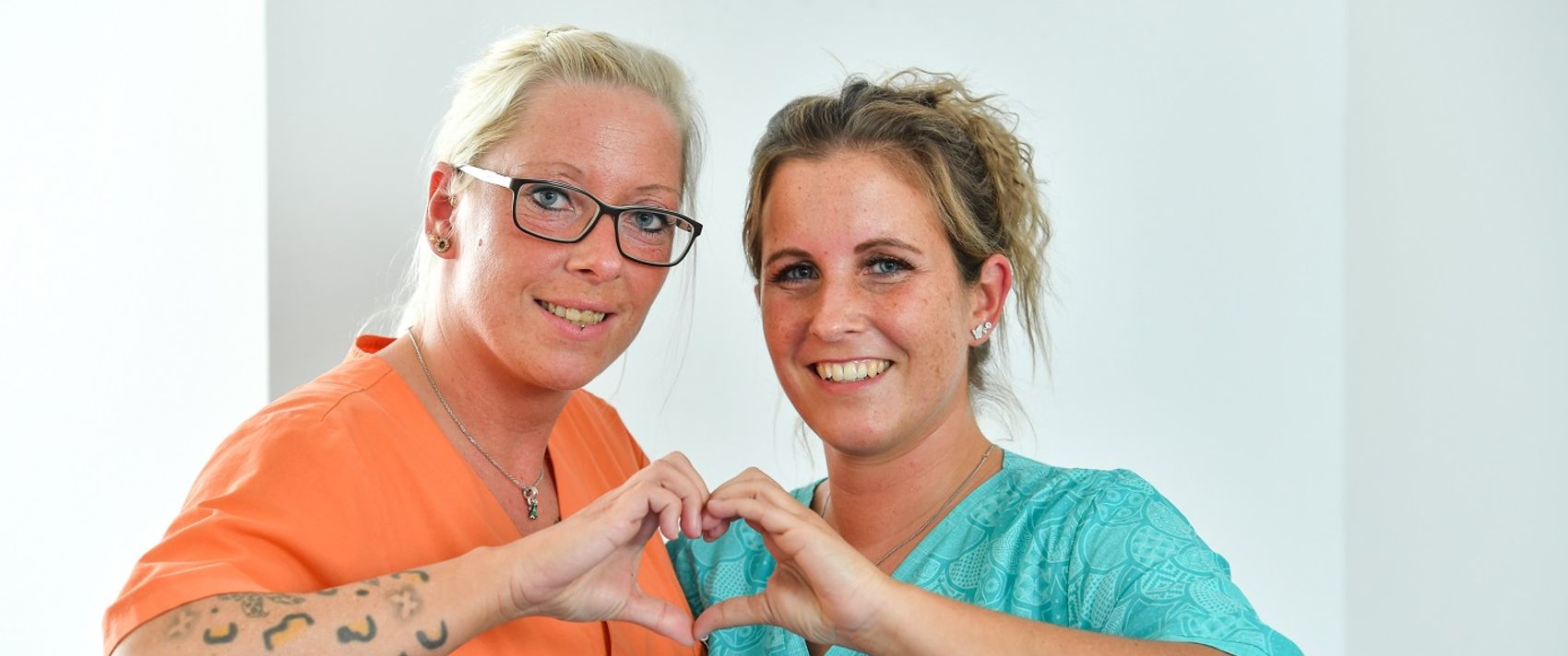 Zwei Frauen formen gemeinsam mit den Händen ein Herz und lächeln dabei in die Kamera