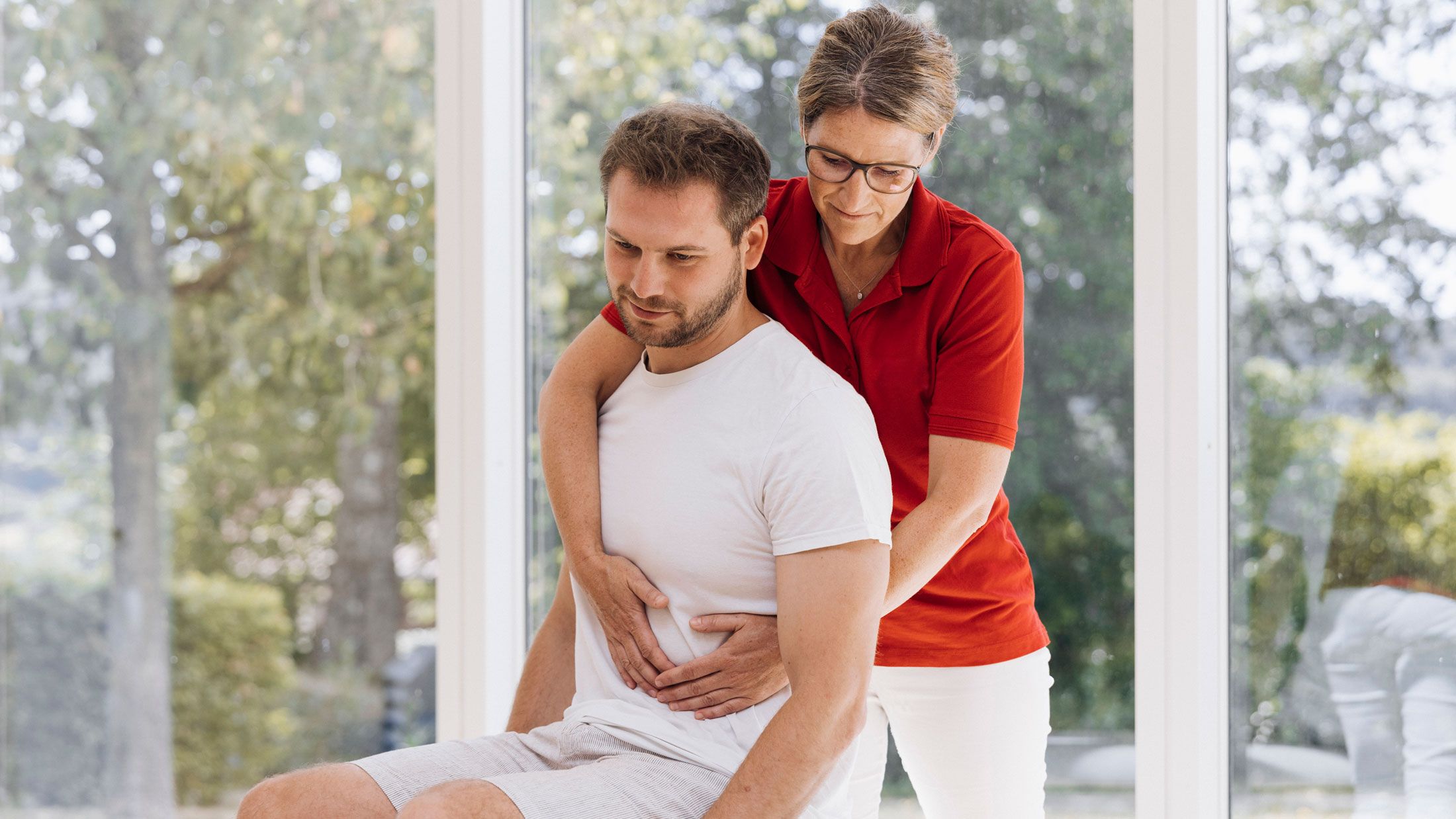 Eine Physiotherapeutin behandelt einen jungen Mann. Im Hintergrund ist eine Glasfront mit Blick in die Natur