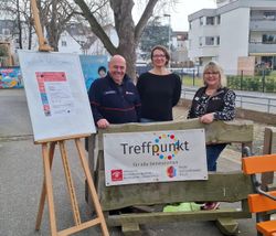 Ein Malteser-Ehrenamtler steht mit zwei Kooperationspartnerinnen im Freien an einem Schild mit dem Titel "Treffpunkt"