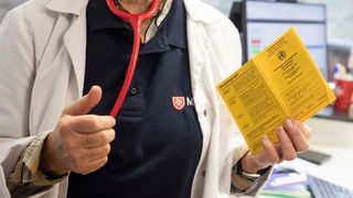 Nahaufnehme eines Menschen in weißem Kittel und Malteser Shirt, der einen Impfpass hochhält. 