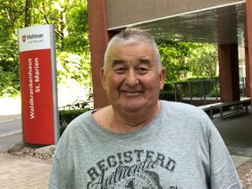 Älterer Mann in grauem T-Shirt steht vor dem Eingang einer Klinik mit rotem Hinweis-schild.