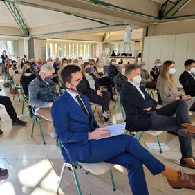 Personen sitzen mit Abstand und teils mit Maske bei einer Veranstaltung in einem Saal.