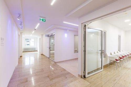 Flur mit Glastür zum Wartebereich und Stuhlreihe entlang der rechten Wand.