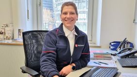 Eine Frau am Schreibtisch sitzend lächelt in die Kmaera.