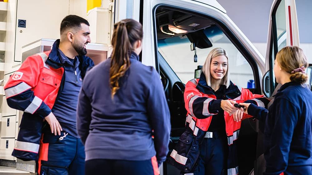 Vier Freiwillige des Malteser Rettungsdienst stehen vor einem Rettungswagen und lächeln sich gegenseitig an.