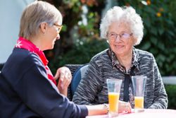 Zu sehen sind zwei Damen draußen an einem Tisch. Links eine Ehrenamtliche, die ein rotes Malteser Halstuch trägt und die ältere Dame berät, die rechts sitz. Die erste Dame ist im Profil zu sehen. Die ältere Dame rechts frontal.