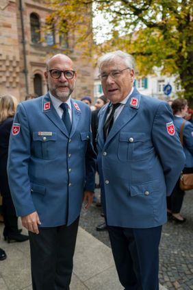 Zwei ältere Männer in blauen Uniformjacken stehen im Freien vor einer Menschenmenge.