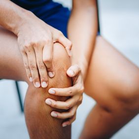 Person hält sich mit beiden Händen schmerzhaft das Knie, sitzend und in sportlicher Klei-dung.