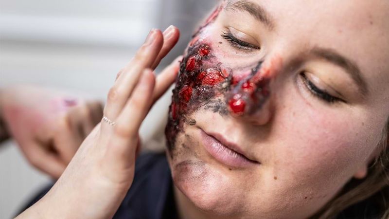 Die Hände einer Frau, die eine künstliche Brandwunde auf das Gesicht einer anderen Frau auftragen. 