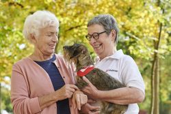 Zwei Frauen stehen vor herbstlich gefärbten Bäumen. Eine Frau trägt ein weißes Malteser-Poloshirt und hält einen kleinen Hund mit rotem Malteser-Halstuch im Arm.