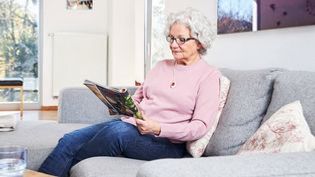 Eine ältere Dame sitzt auf einem Sofa und liest eine Zeitschrift.