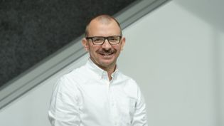 Porträt einer Person mit Brille und weißem Hemd vor modernem, hellen Hintergrund.
