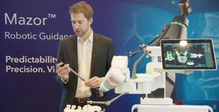 Person demonstriert den Operationsroboter Mazor X mit Roboterarm, Bildschirm und eingesetztem Instrument.