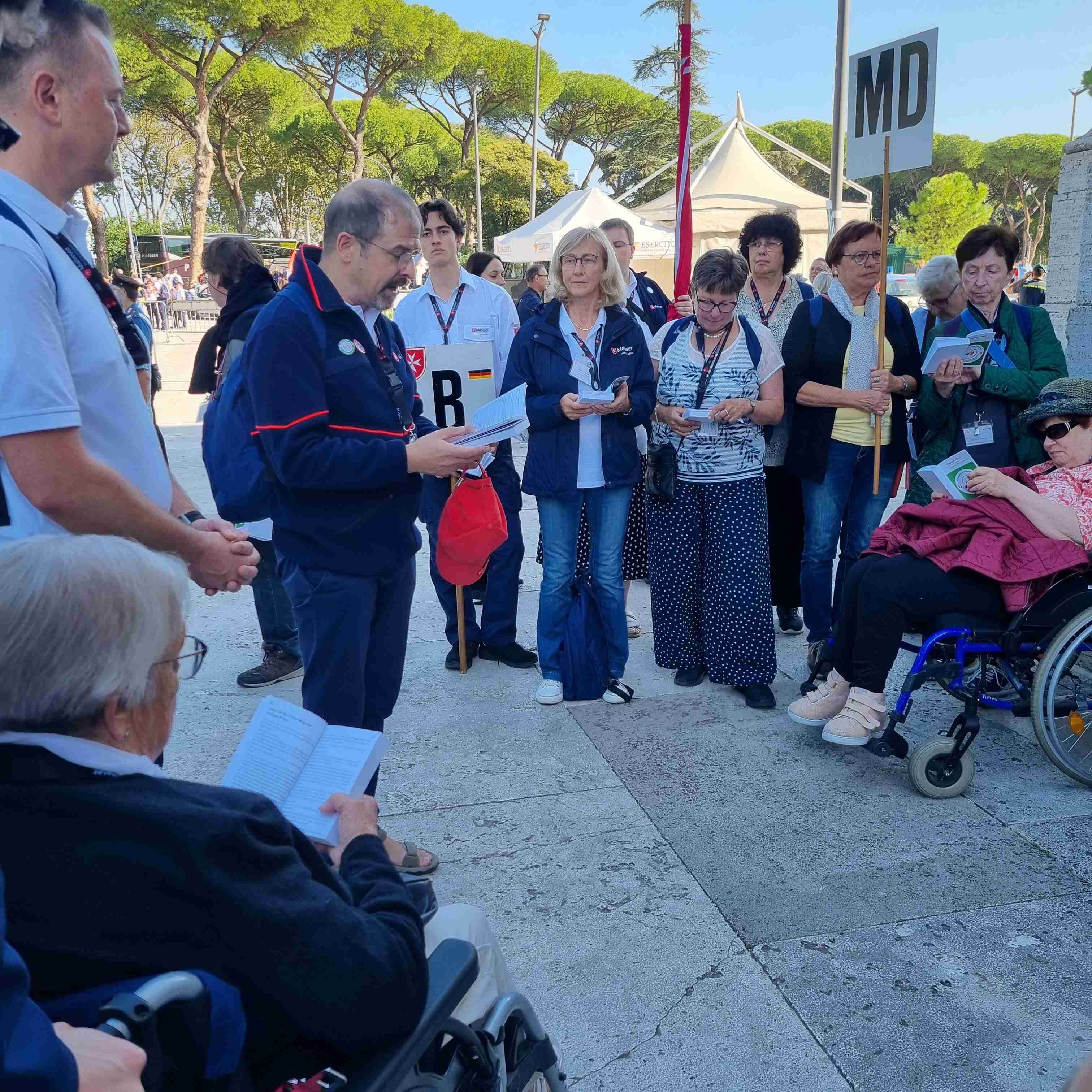 Gruppe von Menschen steht im Freien auf gepflastertem Platz, einige sitzen in Rollstühlen, im Hintergrund Bäume und Zelte.