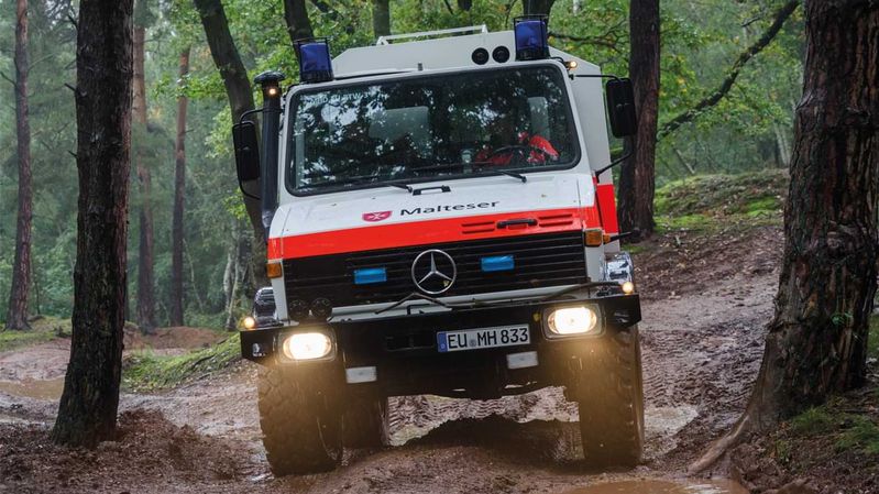 Ein Malteser Unimog fährt bei Regen durch ein hügeliges Waldgebiet. 