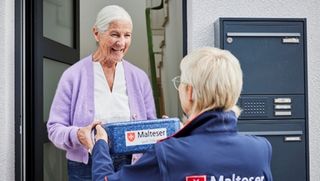 Eine Dame übernimmt an der Haustüre die Menü-Box von einer Malteser Helferin