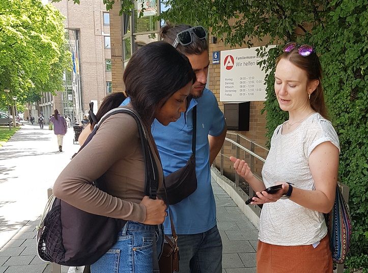Drei Personen schauen gemeinsam auf ein Handy, im Hintergrund das Arbeitsamt München