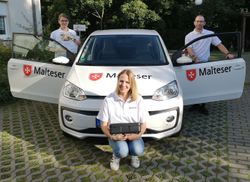 Drei Mitarbeitende des Malteser Hilfsdienst‑Menüservice stehen beziehungsweise knien vor einem weißen Dienstfahrzeug mit Malteser‑Logo. Links hält eine Frau ein Prospekt in der Hand, rechts steht ein Mann mit geöffnetem Fahrertür, vorne kniet eine Frau und hält eine isolierte Essensbox.
