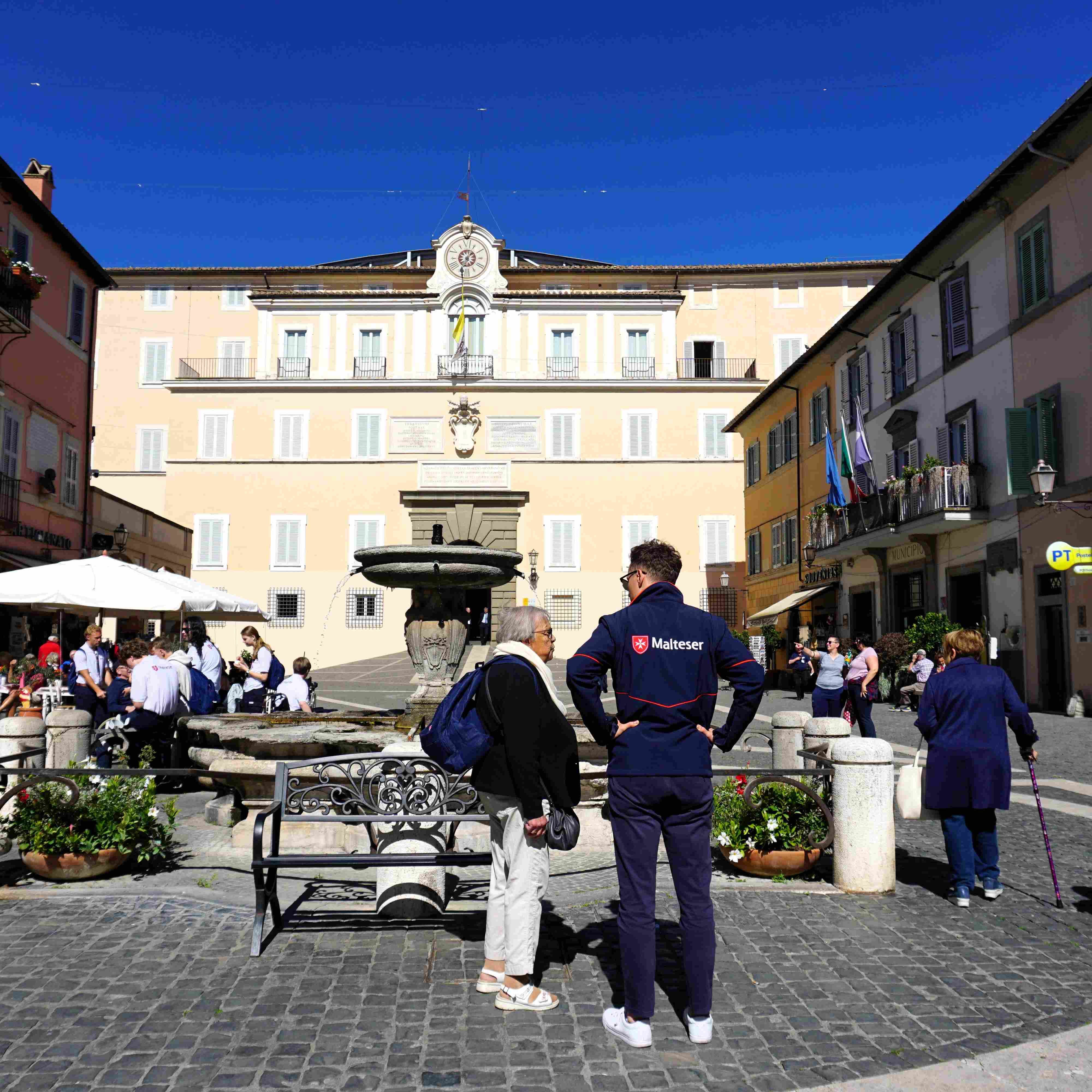 Platz in Castel Gandolfo mit Brunnen, umgeben von mehrstöckigen Gebäuden, Menschen stehen und sitzen bei sonnigem Himmel.
