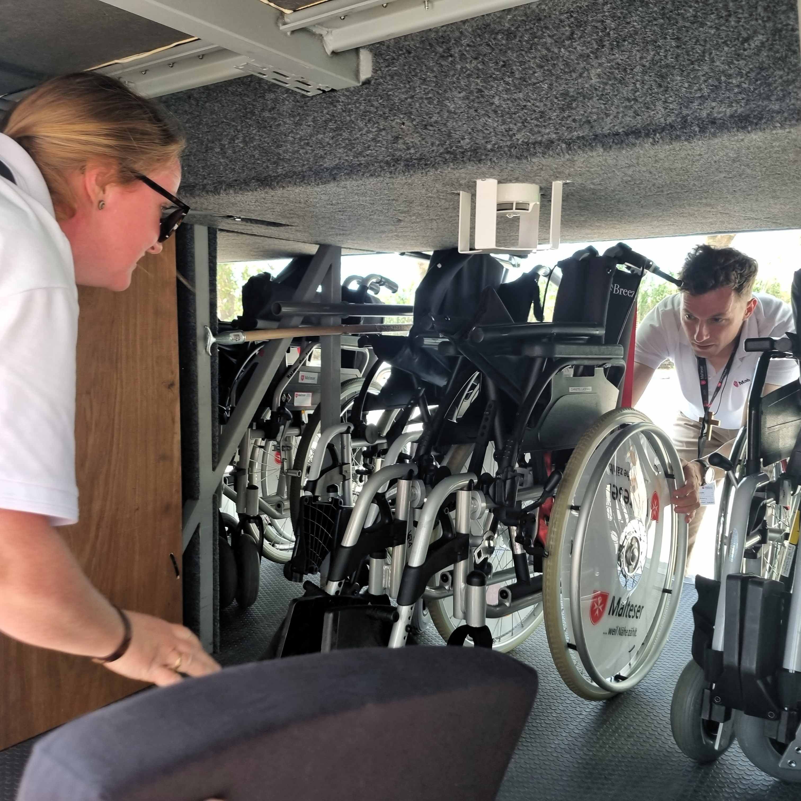 Person in weißem Hemd beugt sich über einen Rollstuhl, im Hintergrund weitere Rollstühle in einem Raum.