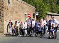 Gruppe von Menschen, darunter mehrere im Rollstuhl, geht eine Straße entlang neben einer hohen Steinmauer mit Verkehrsschildern, darunter ein rotes Einfahrtverbotsschild.