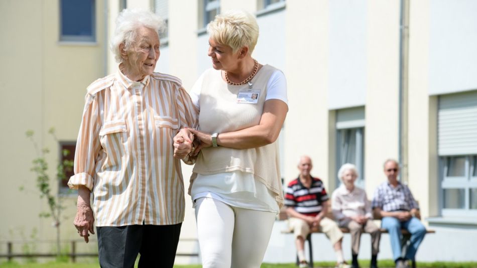 Eine ältere Person wird auf dem Außengelände einer Pflegeeinrichtung von einer Betreuungskraft beim Gehen begleitet, während weitere Bewohnerinnen und Bewohner im Hintergrund sitzen.