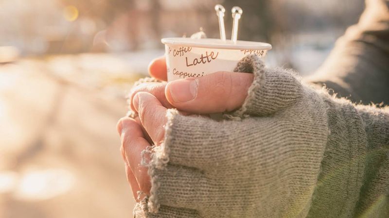 Jemand hält einen Kaffeebecher mit fingerlosen grauen Handschuhen.