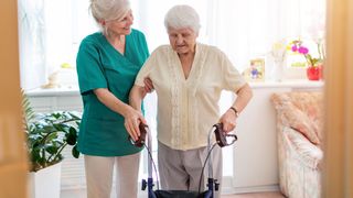 Eine Pflegerin hilft einer älteren Dame mit Rollator beim gehen