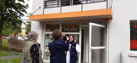 Drei Personen stehen vor dem Eingang eines Treffpunkts der Malteser: Eine Frau mit Kopftuch in einer Malteser‑Weste wird von zwei Menschen angesprochen bzw. interviewt. Im Hintergrund ist ein Gebäude mit geöffneten Türen und ein orangefarbener Balkenvorsprung über dem Eingang zu sehen.