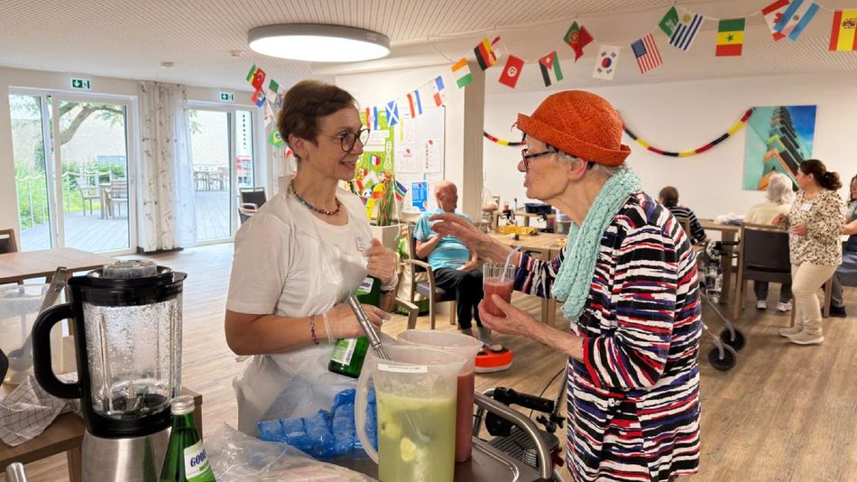 Eine ältere Frau mit Hut unterhält sich mit einer jüngeren Frau in weißer Arbeitskleidung an einer Getränkeausgabe. 