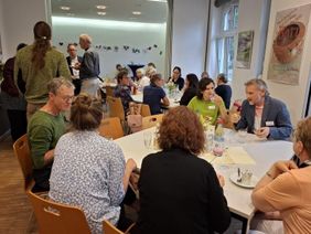 Sechs Personen sitzen an einem Tisch, essen und trinken. Im Hintergrund stehen Leute, die sich unterhalten. 